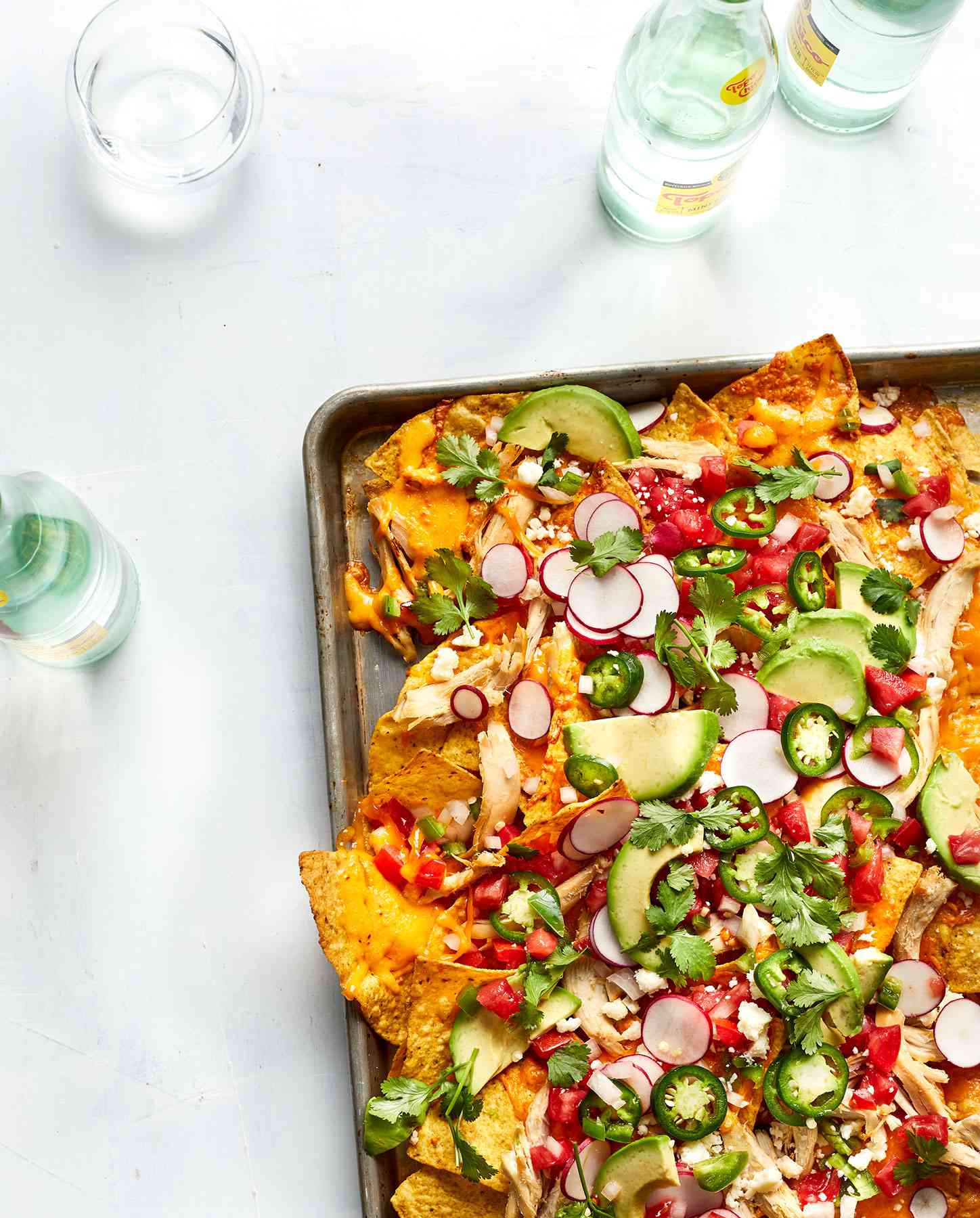 Cheesy Chicken Nachos With Avocado and Radish on a Baking Sheet, Surrounded by Glasses and Bottles of Water