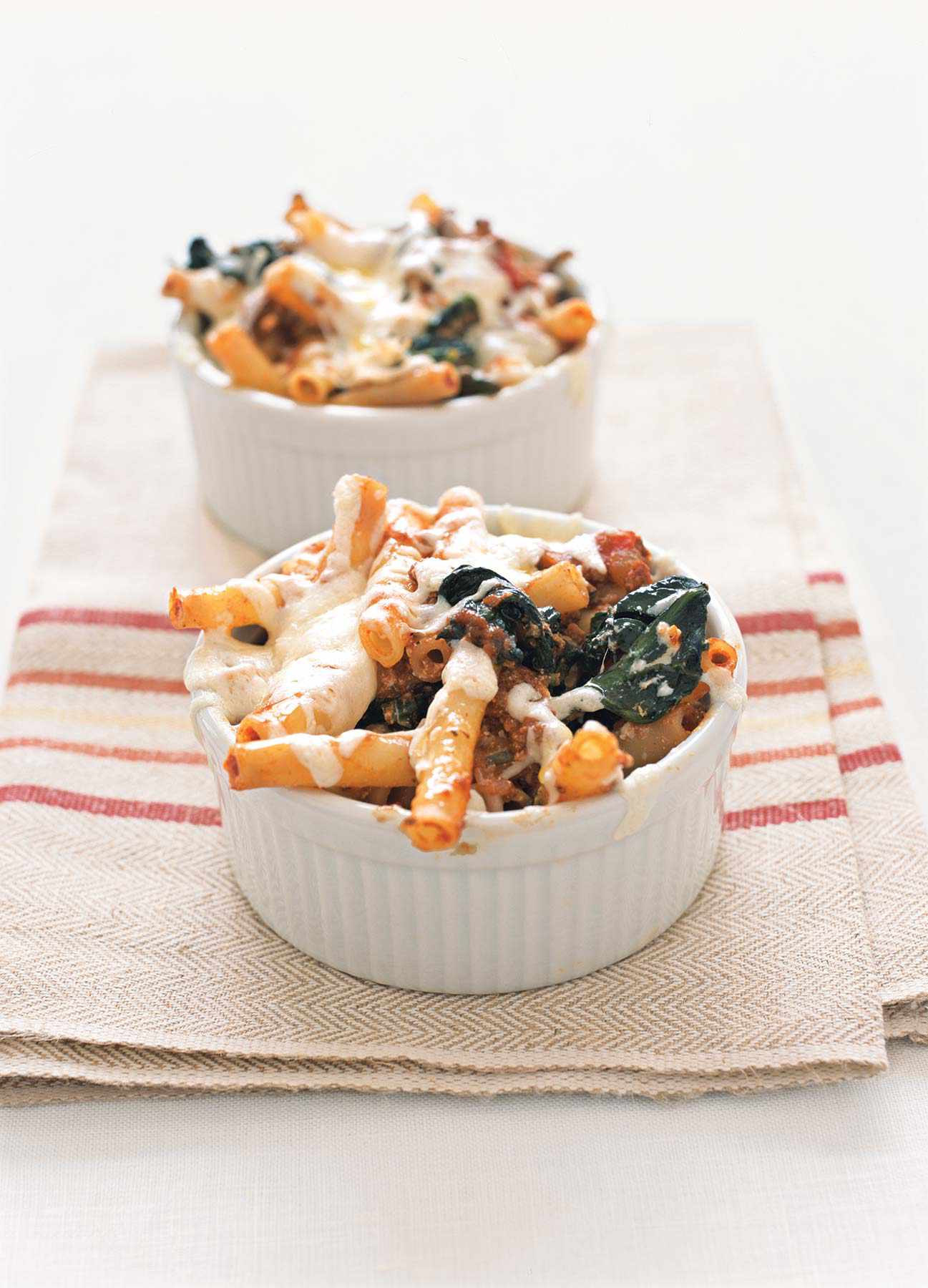 Two white ramekin bowls of lasagna-style baked ziti sitting on a striped kitchen towel.