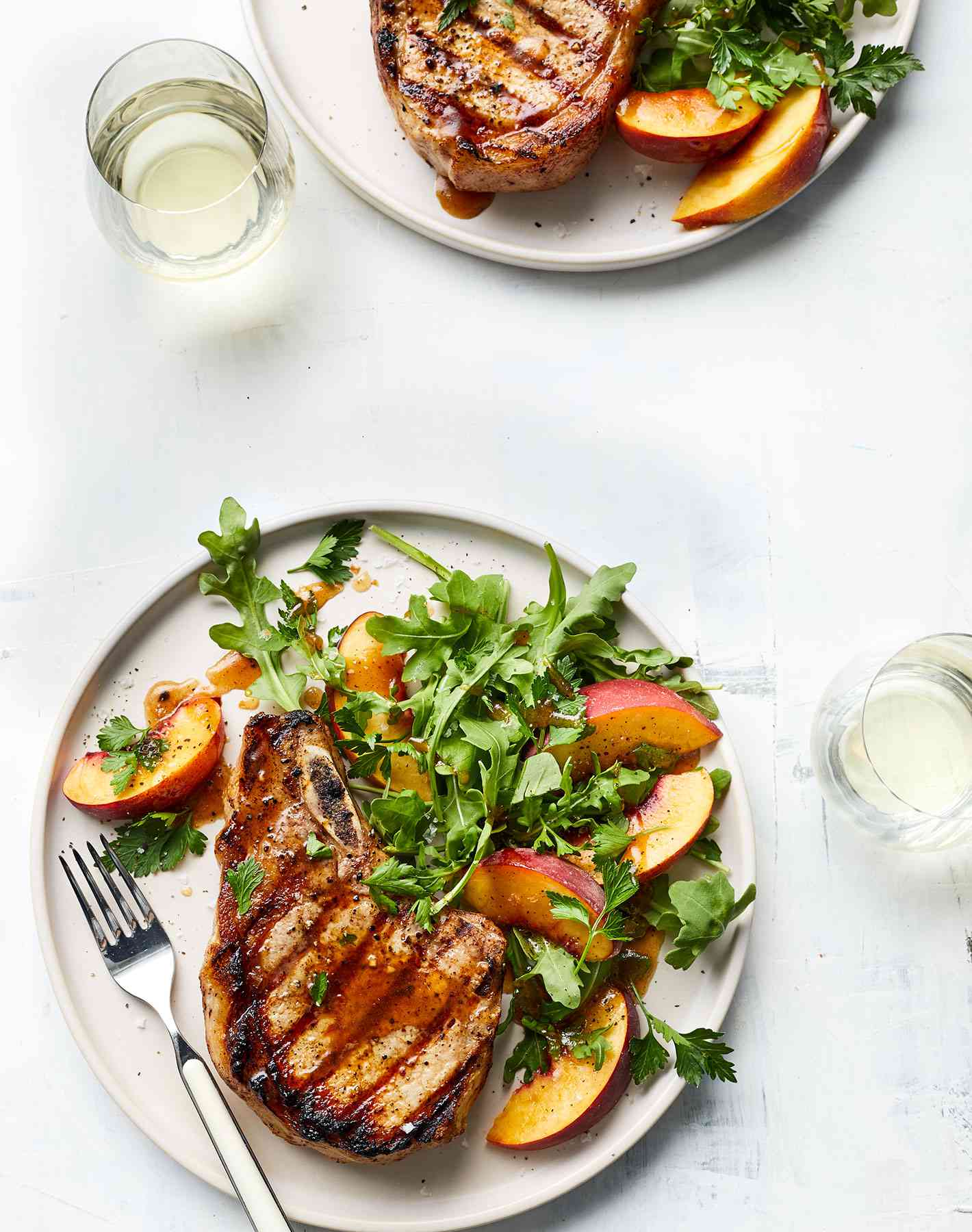 Grilled Pork Chops with Peach Arugula Salad Served on Two Circular White Plates With a Metal Fork
