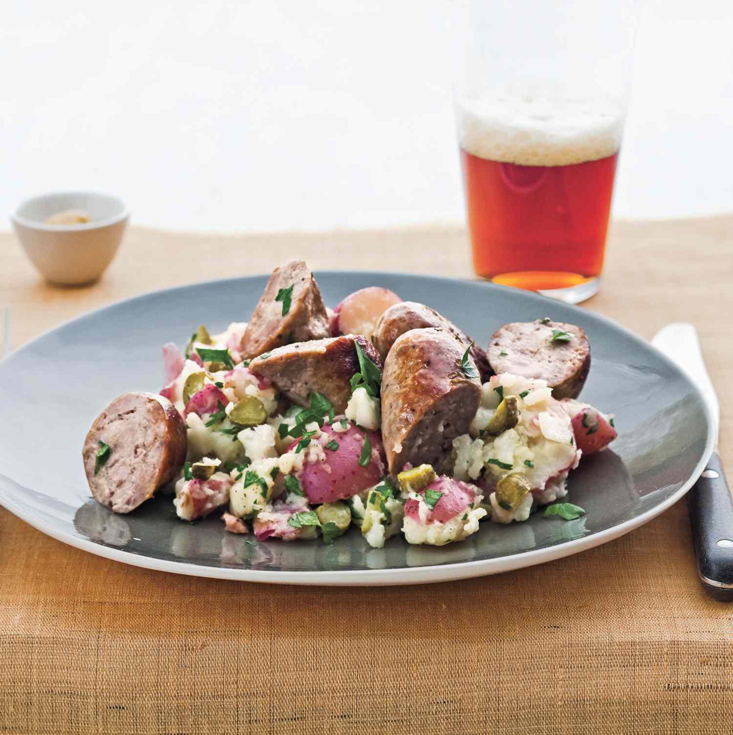 Sausage With Smashed Potatoes and Cornichons on a Gray Plate, Glass of Beer in the Background