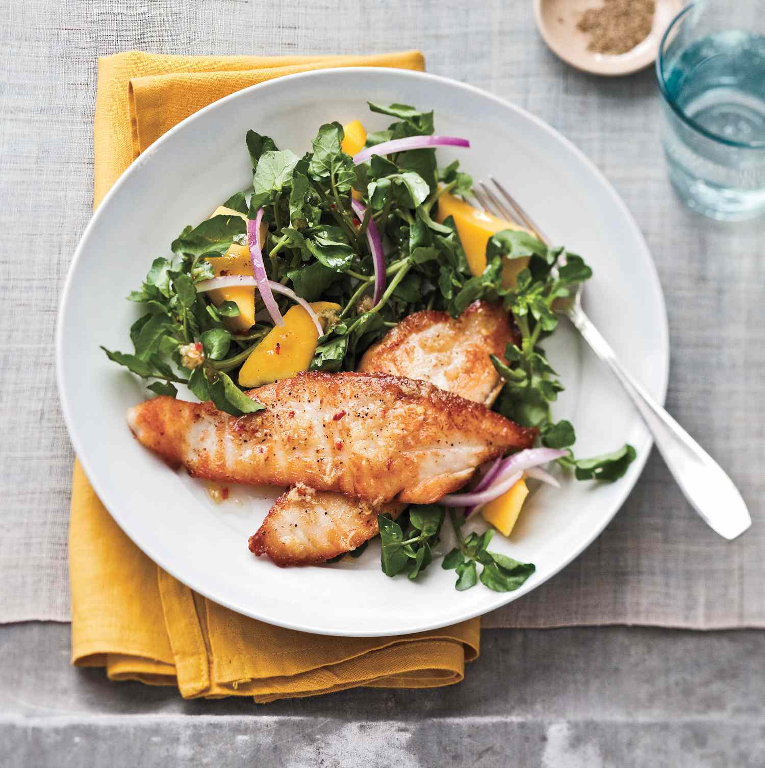 Seared Tilapia Served With Watercress Mango Salad on a White Plate Sitting on a Yellow Cloth Napkin With a Metal Fork for Eating