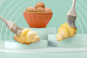two croissants are being brushed with egg wash, an orange bowl with four brown eggs sits in the background